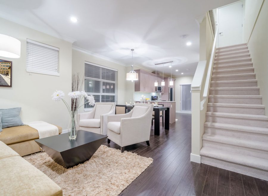 A stylishly finished basement with a neutral color scheme, featuring a beige sectional sofa, two accent chairs, and a sleek black coffee table on a plush rug. The space includes a carpeted staircase leading upward, dark hardwood floors, and a dining area visible in the background.