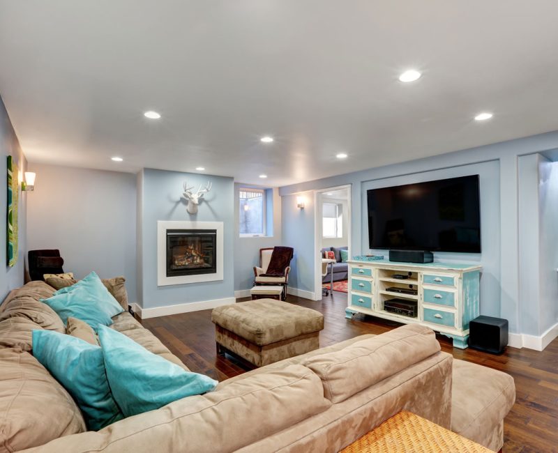 A comfortable finished basement featuring a large sectional sofa with turquoise throw pillows, a fireplace with a mounted deer head above, and a flat-screen TV on a light blue media console. The room has hardwood flooring, recessed lighting, and a staircase leading to the upper level.