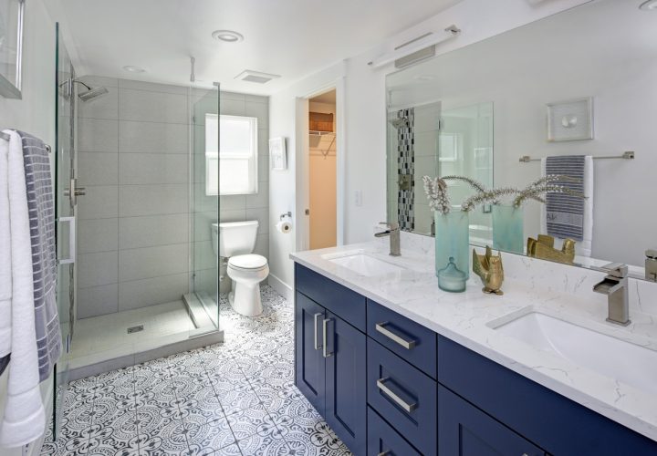 A spacious, modern bathroom with a double vanity featuring navy blue cabinets and a white marble countertop. The room includes a walk-in shower with glass doors, a toilet, patterned black-and-white floor tiles, and a large mirror with decorative accents like vases and towels.