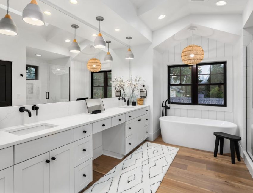A bright, airy bathroom featuring a long white vanity with dual sinks, black fixtures, and a large mirror. The room includes a freestanding bathtub near a window with dark trim, a glass shower enclosure, wooden flooring, and stylish pendant lighting with woven shades.