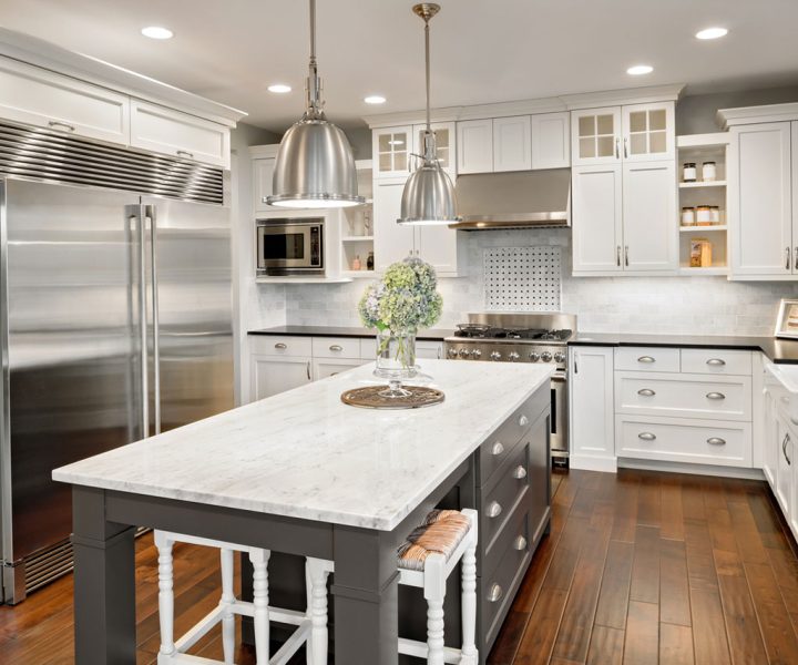 A sophisticated kitchen with white shaker cabinets, a large marble-topped island with dark base and white barstools, and stainless steel appliances including a built-in refrigerator and range hood. The room features hardwood flooring, pendant lights, and a vase of flowers on the island.