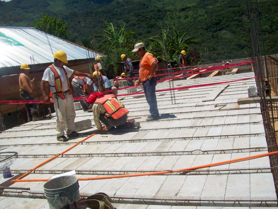 A group of construction workers in hard hats and safety vests are installing concrete roof slabs on a building under construction. The site features metal rebar grids, orange hoses, and red caution tape, with lush green hills and tropical vegetation in the background.